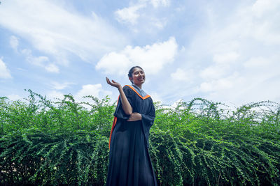 Low angle portrait of woman wearing graduation gown standing against sky