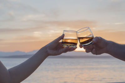 Cropped hands of couple toasting drinks at beach during sunset