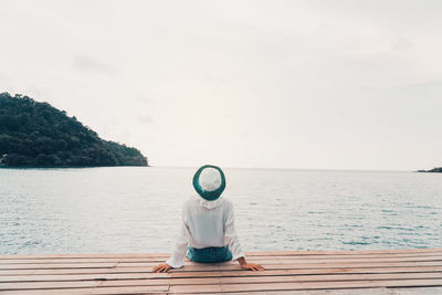 Rear view of girl looking at sea while sitting on pier against sky