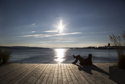 Full length of silhouette man relaxing on boardwalk by lake against sky