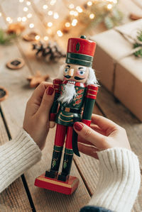 Close-up photo of woman holding wooden nutcracker soldier in front of lights and christmas present