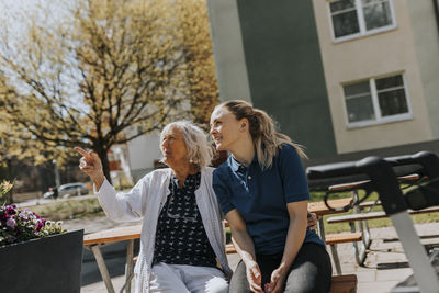 Senior woman gesturing while sitting with female caregiver on bench