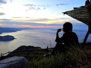 Silhouette woman sitting on cliff by river at sunset