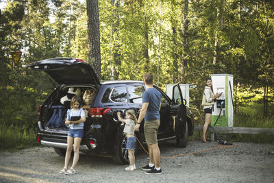 Portrait of mother, father and two daughters standing by car at electric vehicle charging station