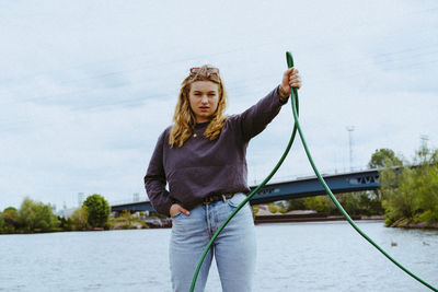 Portrait of confident young blond woman holding garden hose against sky