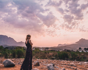 Woman standing on dirt road against sky during sunset