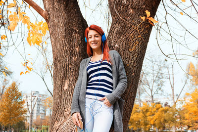 Portrait of woman listening to music while standing against tree trunk during autumn