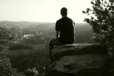 Rear view of man sitting on cliff overlooking landscape