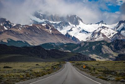 Empty road amidst snowcapped mountains against cloudy sky