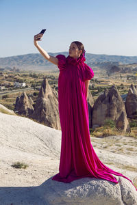 Girl in a big long purple dress stands on a rock in cappadocia and takes a selfie on the phone