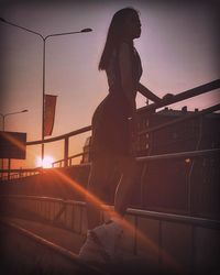 Woman standing on railing against sky during sunset