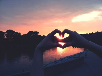 Woman's hands in shape of heart against river at sunset