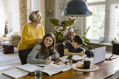 Smiling grandmother with grandchildren doing homework while sitting at table in home