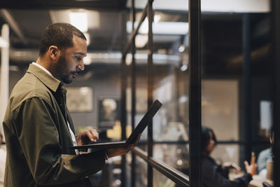 Side view of dedicated businessman using laptop while standing in illuminated office