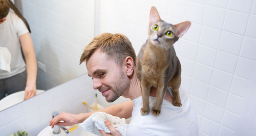 A young smiling blond man is washing his face with a blue abyssinian cat on his shoulders.domestic