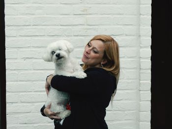 Smiling woman carrying dog while standing against wall