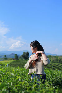 Contemplating woman adjusting hair while standing amidst tea plantation field against sky