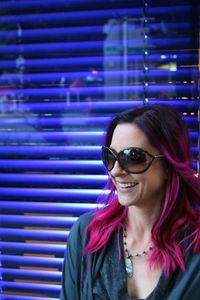 Smiling young woman with dyed pink hair against blue blinds