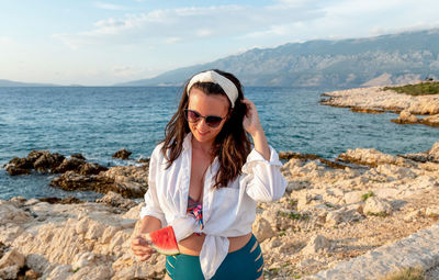 Young woman in bikini and white shirt holding piece of watermelon on beach.