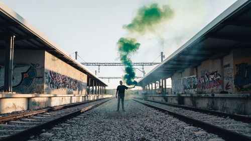 Young man holding smoke bomb while standing on railroad tracks against sky