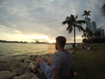 Man sitting on shore against cloudy sky during sunset