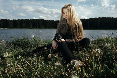 Woman sitting with dog by lake against cloudy sky