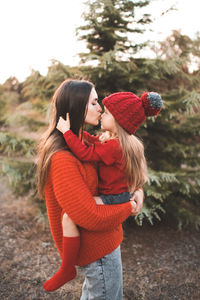 Young woman mom spend time with daughter kid girl wear red knit sweater and hat over tree outdoor. 