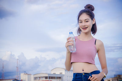 Portrait of smiling young woman with water bottle standing against cloudy sky
