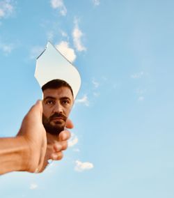 Cropped hand of man holding broken mirror against sky