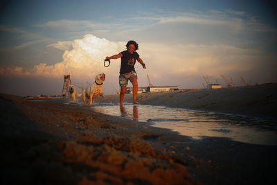 Woman with dogs running on shore at beach against sky during sunset