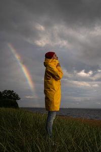Male in yellow raincoat wear red hat standing on the beach in rainy weather.