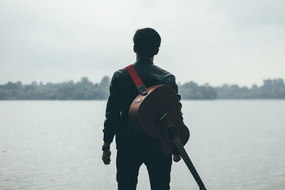 Rear view of man with guitar standing by lake against sky