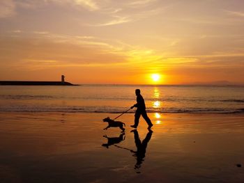 Silhouette man with dog walking at beach against sky during sunset