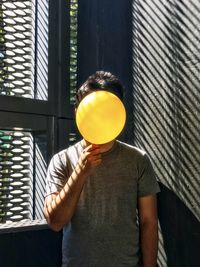 Midsection of man holding yellow balloon against window and wall.
