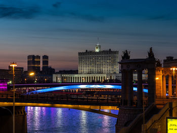 Illuminated bridge over river by buildings against sky at dusk