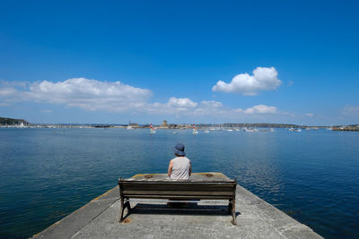 Rear view of woman looking at sea while sitting on bench against blue sky during sunny day