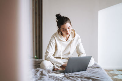 Beautiful smiling woman with dark long hair in casual using laptop sitting on bed at home