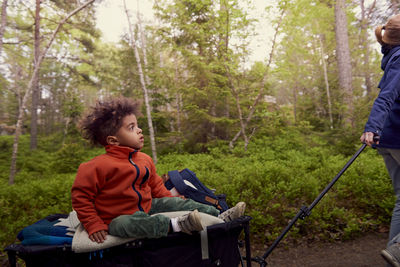 Mother pulling boy sitting on camping cart against trees in forest