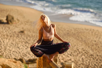Young woman meditating while sitting on rock at beach against sky