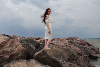 Side view of young woman standing on rocks at beach against sky