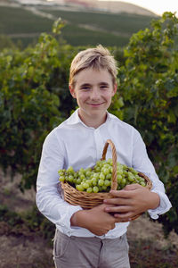Teenage boy in a white shirt stands in a vineyard at sunset and holds a basket of green grapes