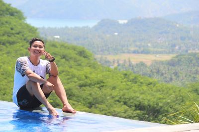 Portrait of smiling young man sitting by swimming pool against mountain
