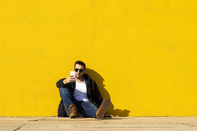 Young bearded man with sunglasses sitting on yellow wall, using phone