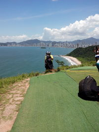 Rear view of people sitting on grass by sea against sky