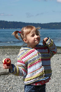 Side view of cute little girl standing at beach, eating apple, enjoying beautiful day