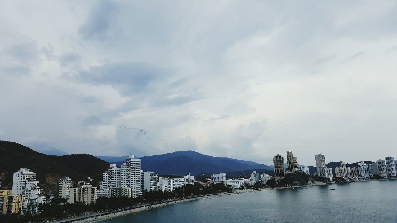 water, waterfront, architecture, built structure, city, mountain, building exterior, sky, cityscape, mountain range, sea, cloud - sky, development, river, skyscraper, city life, scenics, residential district, tall - high, outdoors, distant, day, cloudy, modern, tranquil scene, in front of, coastline, office building, riverbank, tranquility