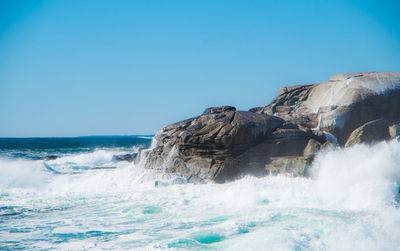 Waves splashing on rock formation against clear blue sky