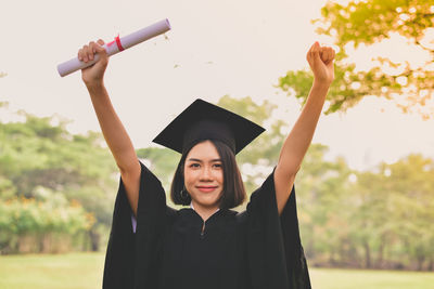 Portrait of young woman in graduation gown with arms raised holding certificate while standing at park