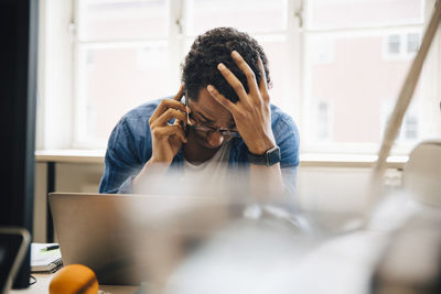 Frustrated male computer hacker with hand in hair talking on smart phone while using laptop in creative office