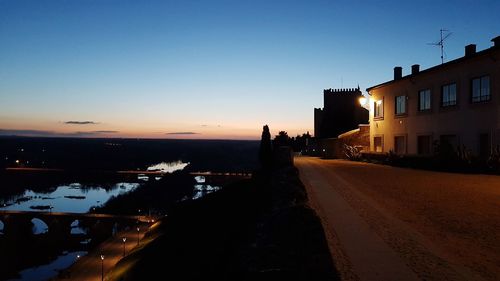 Road by illuminated buildings against sky at sunset